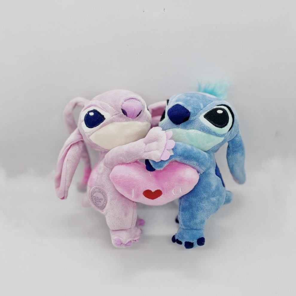 Stich in blue and Angel in pink cuddling each other. They have a pink heart between their arms. The two plushies look like they are in love. The cuddly toys are made of cotton.
