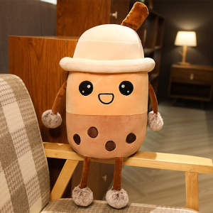 Bubble tea plush with a hat, she is beige and brown, and is sitting on the wooden armrest of a beige, white and brown checked armchair in a flat with a lamp lit in the background