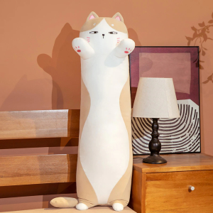 On a bed with a bedside table next to it, with a white bedside lamp and a frame behind it, is a white and brown cat pillow, standing on its tiny hind legs