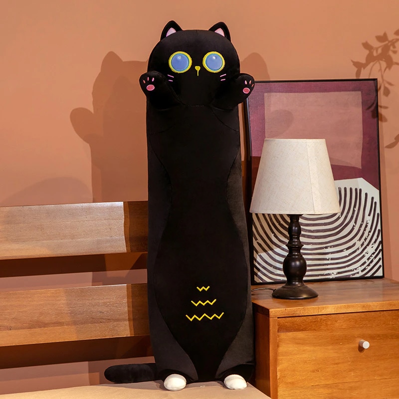 On a bed next to which is a bedside table with a white bedside lamp and a frame behind it, there is a plush black cat pillow, it stands on its tiny back legs