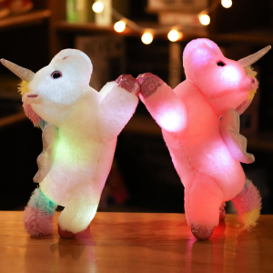 On a wooden table, 2 unicorn plushies, one white and one pink are raised on their back legs and are holding each other by the front one and their bodies are illuminated