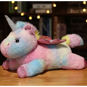 On a wooden table a multicoloured unicorn plush is lying on its stomach with shelves and lights in the background