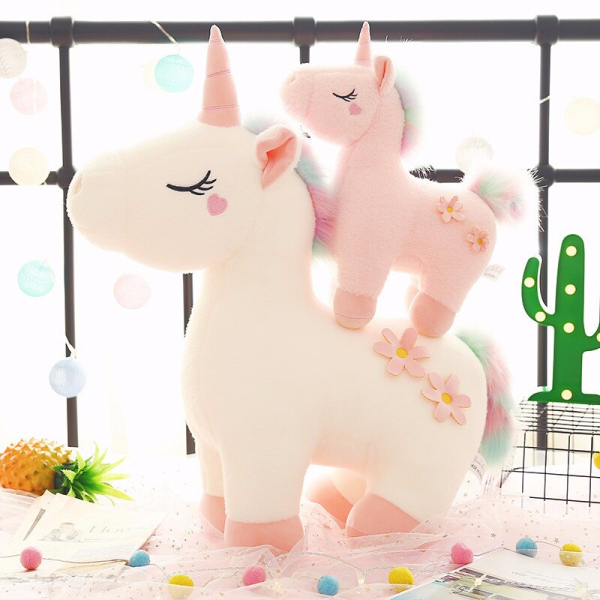 On a terrace, in front of an iron balustrade, 2 unicorn plush toys, one big white one and the other small pink one which is on the back of the first one, next to a small fake cactus