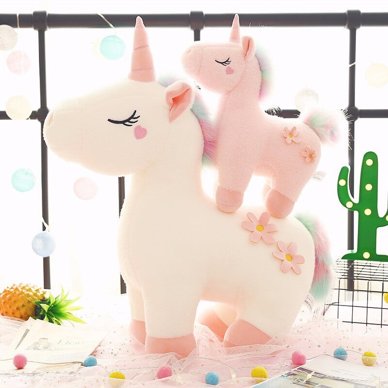 On a terrace, in front of an iron balustrade, 2 unicorn plush toys, one big white one and the other small pink one which is on the back of the first one, next to a small fake cactus
