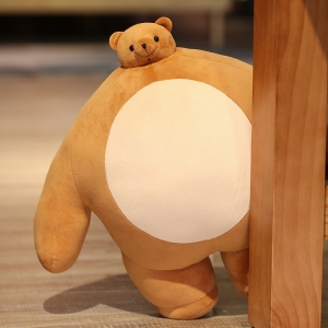 Brown plush bear cushion with a beige belly in front of the foot of a piece of wooden furniture on a wooden floor