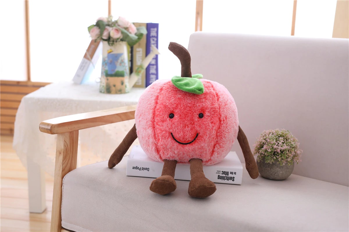 Smiling pink cherry plush on a book and a grey sofa next to a small plant and a coffee table with books in the background