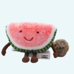 Pink and green watermelon shaped plush sitting on a book on a grey sofa with a table with books in the background