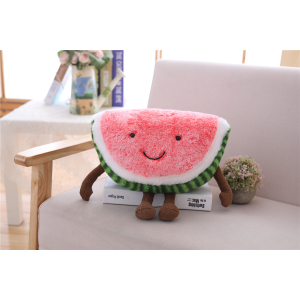 Pink and green watermelon shaped plush sitting on a book on a grey sofa with a table with books in the background