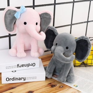 Two elephant cuddly toys, one pink and one grey. The pink one is placed on two white and black books which are placed on a table, the grey one is placed next to it on the wooden table. In the background white tiles