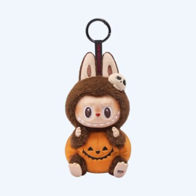 Brown Labubu plush dressed as a pumpkin with a small felt skull on its head and a keychain loop, on a neutral background